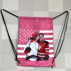 May include: Pink drawstring bag featuring a graphic of two hockey players, one kissing the other. The bag has a design resembling the American flag with stars and stripes. Black drawstrings and gold eyelets complete the design.