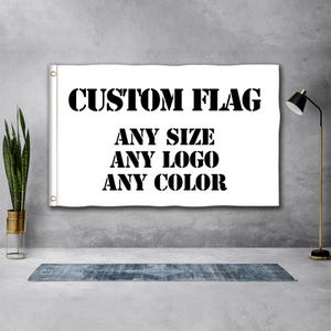 以下が含まれることがあります： 「CUSTOM FLAG、ANY SIZE、ANY LOGO、ANY COLOR」と書かれた黒い文字が入った白いカスタムフラッグ。旗は灰色の壁に掛けられています。旗には吊り下げ用のグロメットが付いています。左側には、金色の鉢に入った背の高い植物があります。