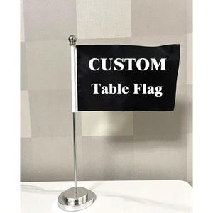 May include: A black custom table flag with white text. The flag is mounted on a silver pole with a round base. The text on the flag reads "CUSTOM Table Flag". The flag is displayed on a white surface.