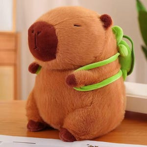 Könnte beinhalten: Ein braunes Capybara-Plüschtier mit einer dunkelbraunen Nase und Füßen. Es hat einen kleinen grünen Rucksack mit grünen Trägern. Das Kuscheltier ist ideal für Kinder oder Sammler.
