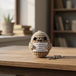 Peut inclure: Un porte-clés en forme de pomme de terre crochetée, avec des yeux en boutons noirs et un petit sourire. La pomme de terre tient une pancarte avec l'inscription : « Life was all out of lemons, so here's a potato instead ». Un porte-clés argenté est posé sur une surface en bois.