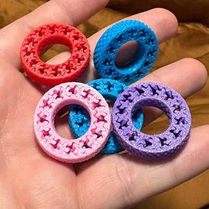 May include: A collection of colorful, 3D-printed rings in red, blue, pink, and purple. Each ring features a repeating star-shaped pattern around its circumference. The rings are held in a hand, showcasing their vibrant colors and intricate design.
