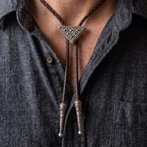 May include: A bolo tie featuring a dark brown braided leather cord and a triangular silver-toned metal slide with a geometric embossed pattern. The cord ends are finished with matching silver-toned metal tips featuring intricate floral engravings.