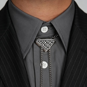 Op de afbeelding: Een zilveren bolo stropdas met een geometrische driehoekige hanger. De hanger heeft een gedetailleerd patroon en hangt aan een ketting. De stropdas wordt gedragen met een grijs overhemd en een zwart krijtstreepjasje.