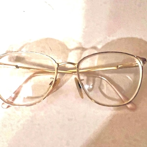 May include: Gold-rimmed eyeglasses with clear lenses. The glasses have a classic, oval shape and a delicate, thin frame. The nose pads are a light beige color. The glasses are resting on a light pink surface.
