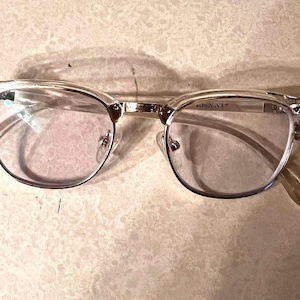 May include: Clear-framed eyeglasses with a silver bridge and arms. The lenses are round and the frame has a classic, retro design. The glasses are resting on a light-colored surface.