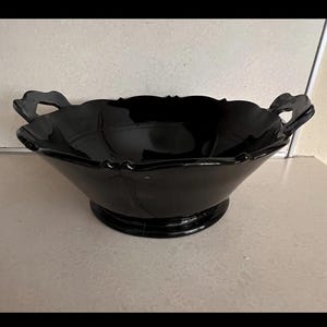 May include: A black glass serving bowl with two handles. The bowl has a scalloped rim and sits on a round base. The bowl is likely used for serving food or as a decorative piece.
