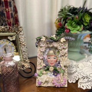 May include: A rectangular decorative frame featuring a central sculpted face with a gold cap, surrounded by floral embellishments, small purple mushrooms, and a miniature fairy figure. The frame is adorned with green velvet ribbon and lavender accents.