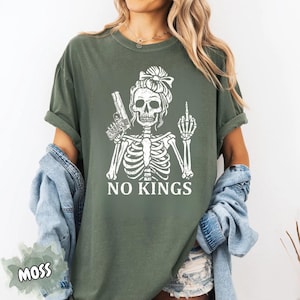 May include: Moss green t-shirt featuring a white skeleton graphic holding a gun and giving the middle finger. The text "NO KINGS" is printed below the graphic. The shirt has short sleeves and a relaxed fit.
