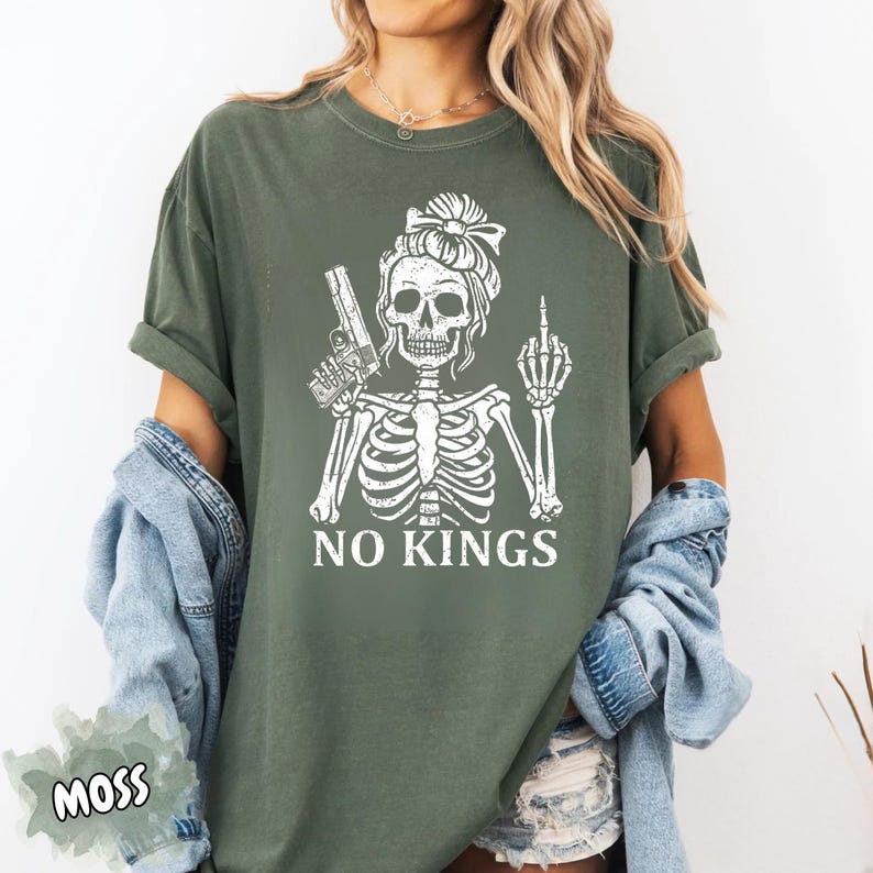 May include: Moss green t-shirt featuring a white skeleton graphic holding a gun and giving the middle finger. The text "NO KINGS" is printed below the graphic. The shirt has short sleeves and a relaxed fit.