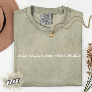 May include: Khaki-colored Comfort Colors t-shirt with the text "looks tough, sleeps with a blankie." A gold star pendant necklace rests on the shirt. The shirt is size XL-TG-EG.