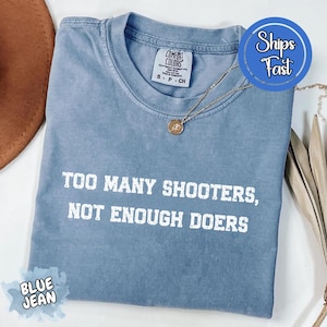 May include: A blue jean Comfort Colors t-shirt with the text "TOO MANY SHOOTERS, NOT ENOUGH DOERS" in white. A gold necklace with a pendant is on the shirt. The shirt is folded and has a "Ships Fast" sticker.