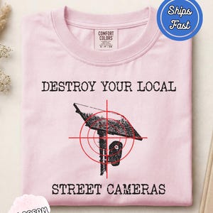 Puede incluir: Una camiseta rosa claro con un gráfico negro de una cámara de calle sobre un poste, centrado en una mira roja. El texto "DESTROY YOUR LOCAL" aparece sobre el gráfico y "STREET CAMERAS" está impreso debajo con una tipografía desgastada.