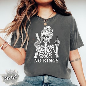 May include: Dark gray t-shirt featuring a white skeleton graphic. The skeleton is holding a gun and giving the middle finger. The text "NO KINGS" is printed below the graphic. The shirt has a relaxed fit.