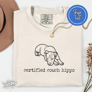 Op de afbeelding: Een ivoorkleurig t-shirt met een zwarte lijntekening van een liggende hond, geplaatst boven de tekst "certified couch hippo" in een typemachine-stijl lettertype. Het t-shirt heeft een Comfort Colors merklabel aan de binnenkant van de hals.