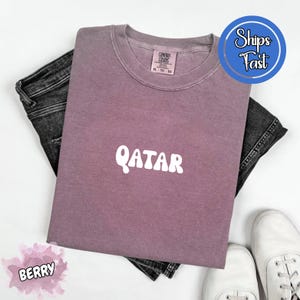 May include: Berry-colored t-shirt with the word "QATAR" in white, paired with dark denim jeans and white sneakers. The shirt has a crew neck and a tag that reads "Comfort Colors".