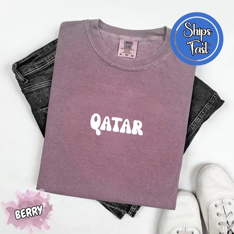 May include: Berry-colored t-shirt with the word "QATAR" in white, paired with dark denim jeans and white sneakers. The shirt has a crew neck and a tag that reads "Comfort Colors".