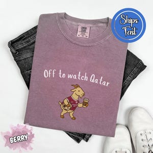 May include: Berry-colored t-shirt with the text "Off to watch Qatar" and a cartoon goat wearing a soccer jersey and holding a coffee cup. The shirt is paired with dark gray jeans and white sneakers.