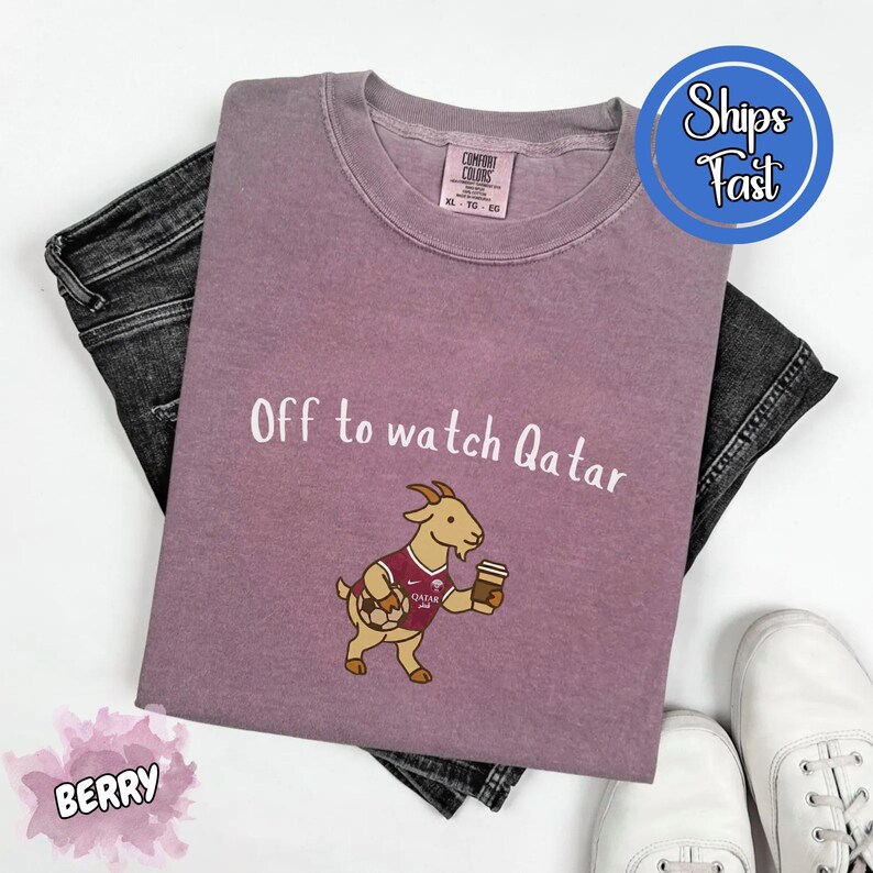May include: Berry-colored t-shirt with the text "Off to watch Qatar" and a cartoon goat wearing a soccer jersey and holding a coffee cup. The shirt is paired with dark gray jeans and white sneakers.