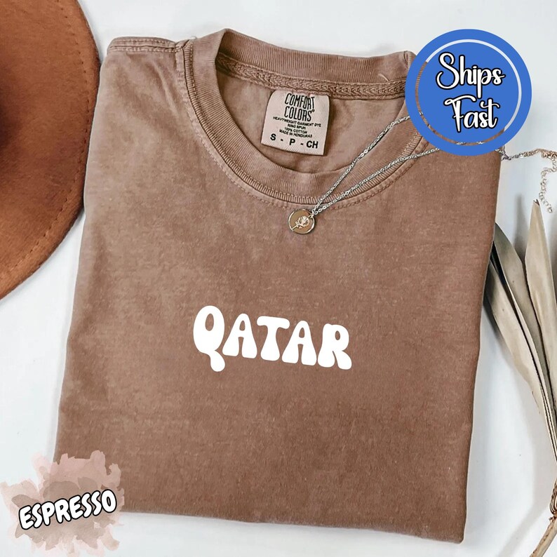 May include: Espresso-colored t-shirt with the word "QATAR" in white, featuring a crew neck and short sleeves. A silver necklace with a pendant is draped on the shirt. The shirt has a "Comfort Colors" tag. A brown hat is partially visible.