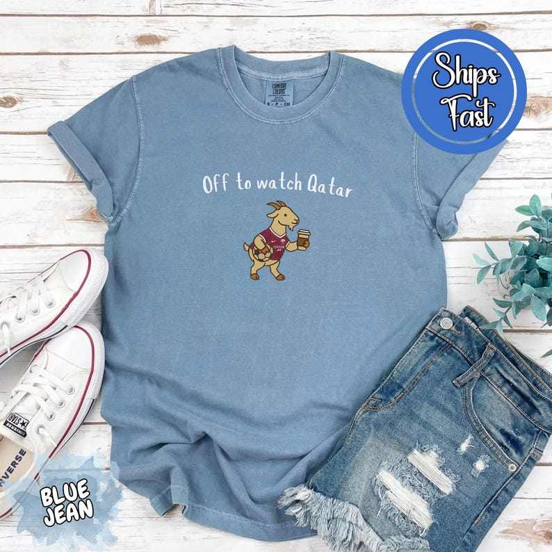 May include: A light blue t-shirt with the text "Off to watch Qatar" and a cartoon goat wearing a soccer jersey and holding a coffee cup. The shirt is paired with blue jean shorts and white sneakers.