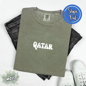 May include: A moss green t-shirt with the word "QATAR" in white, paired with black denim jeans and white sneakers. The shirt has a crew neck and a tag that reads "COMFORT COLORS". A blue circle with "Ships Fast" is in the upper right corner.