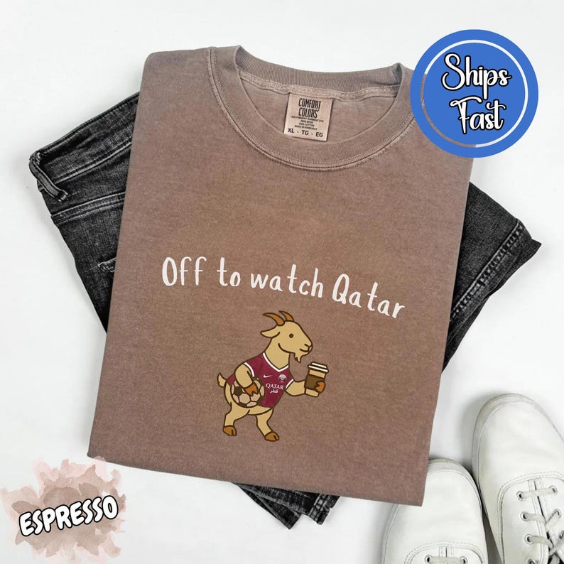 May include: Espresso-colored t-shirt with the text "Off to watch Qatar" and a cartoon goat wearing a soccer jersey. The shirt is paired with dark jeans and white sneakers.