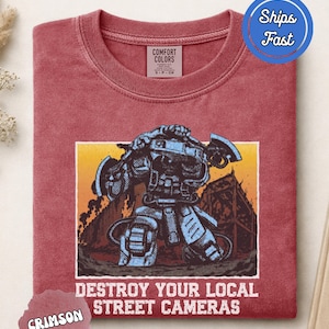 Puede incluir: Una camiseta de cuello redondo color carmesí con un gráfico de una figura robótica sosteniendo hachas sobre un fondo de atardecer. Debajo del gráfico, el texto dice DESTROY YOUR LOCAL STREET CAMERAS. Tiene la etiqueta Comfort Colors en el cuello.