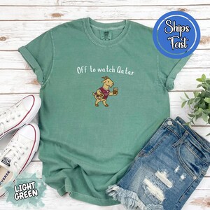 May include: Light green t-shirt with the text "Off to watch Qatar" and a cartoon goat wearing a soccer jersey and holding a coffee cup. The shirt is paired with denim shorts and white sneakers.