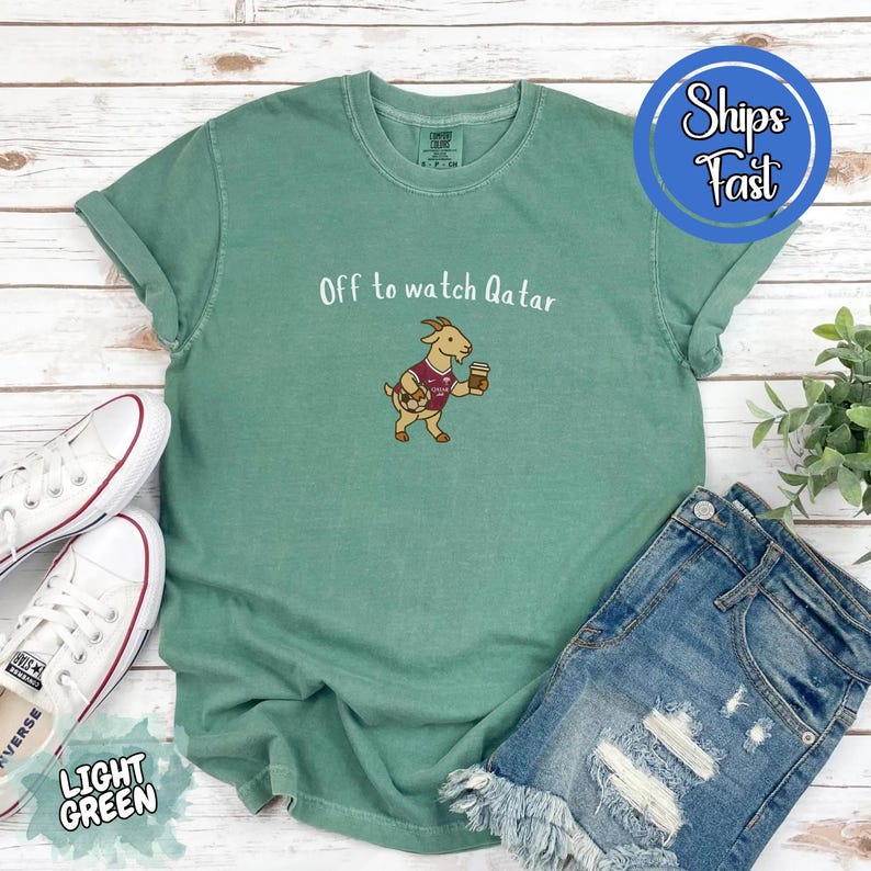 May include: Light green t-shirt with the text "Off to watch Qatar" and a cartoon goat wearing a soccer jersey and holding a coffee cup. The shirt is paired with denim shorts and white sneakers.