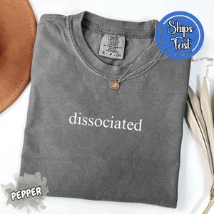 Puede incluir: Una camiseta gris de manga corta con la palabra "dissociated" impresa en blanco. La camiseta tiene cuello redondo y una pequeña etiqueta en la parte superior de la espalda. Un collar plateado con un colgante está sobre la camiseta.