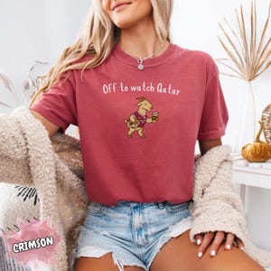 May include: Crimson-colored t-shirt with the text "Off to watch Qatar" and a cartoon goat wearing a jersey and holding a coffee cup. The shirt is paired with denim shorts and a cream-colored sweater.