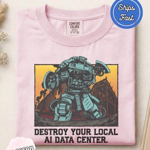 Peut inclure: Un t-shirt rose clair avec une impression graphique représentant un robot mécanique tenant deux haches sur un fond de coucher de soleil jaune et orange. Le texte sous le graphique indique : "DESTROY YOUR LOCAL AI DATA CENTER." Col rond.