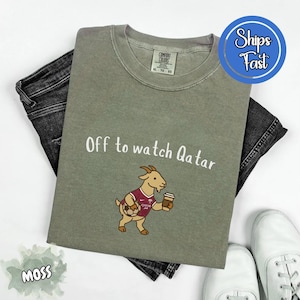 May include: Moss green t-shirt with the text "Off to watch Qatar" and a cartoon goat wearing a maroon jersey and holding a soccer ball and coffee cup. The shirt is laid on top of black jeans and white sneakers.