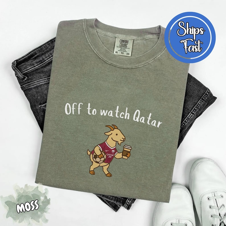 May include: Moss green t-shirt with the text "Off to watch Qatar" and a cartoon goat wearing a maroon jersey and holding a soccer ball and coffee cup. The shirt is laid on top of black jeans and white sneakers.