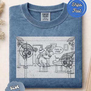 Puede incluir: Una camiseta de color azul vaquero con una ilustración gráfica de un perro con sombrero, sentado a una mesa con una taza, rodeado de cámaras de seguridad y paneles solares, con un bocadillo que contiene el texto THIS IS FINE.