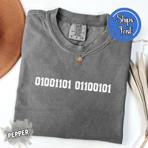 May include: A gray Comfort Colors t-shirt with the binary code "01001101 01100101" printed in white. The shirt has a crew neck and short sleeves. A silver necklace with a pendant is on the shirt.