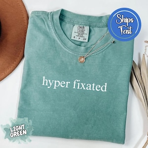 May include: Light green t-shirt with the words "hyper fixated" in white. The shirt has a crew neck and short sleeves. A silver necklace with a small pendant is on the shirt. The shirt is folded and has a "Comfort Colors" tag.