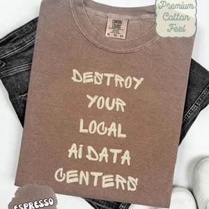 Puede incluir: Una camiseta marrón de cuello redondo con el texto "DESTROY YOUR LOCAL AI DATA CENTERS" impreso en color crema claro. La prenda cuenta con una etiqueta de la marca Comfort Colors en el cuello interior, indicando la talla XL, sobre tela vaquera gris.