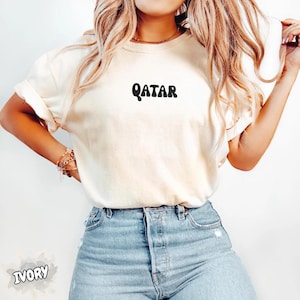 May include: Ivory-colored t-shirt with the word "QATAR" printed in black, featuring a retro font. The shirt is paired with light blue denim jeans. The word "IVORY" is printed at the bottom left corner of the image.