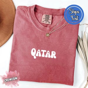 May include: Crimson-colored t-shirt with the word "QATAR" in white, featuring a crew neck and a small tag. A silver necklace with a pendant is draped across the shirt. The shirt is folded on a white surface.