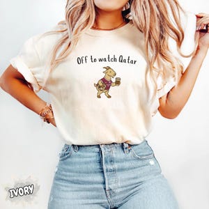May include: Ivory-colored t-shirt with the text "Off to watch Qatar" and a cartoon goat wearing a maroon jersey and holding a coffee cup. The goat is also holding a soccer ball. The shirt is paired with blue jeans.