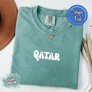 May include: Light green t-shirt with the word "QATAR" in white, rounded letters. The shirt has a crew neck and short sleeves. A silver necklace with a small pendant is on the shirt. The shirt is folded on a surface.