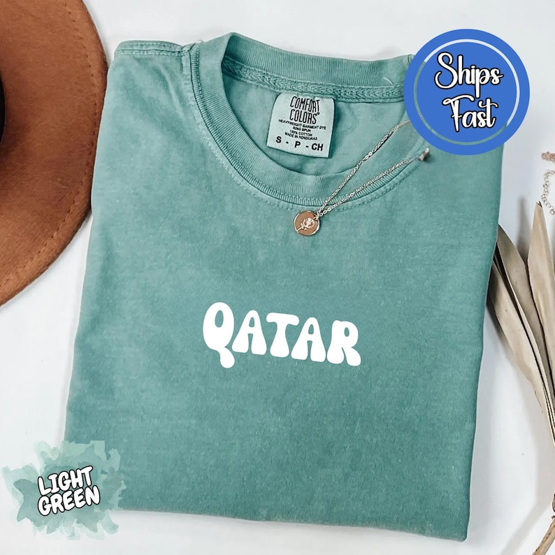 May include: Light green t-shirt with the word "QATAR" in white, rounded letters. The shirt has a crew neck and short sleeves. A silver necklace with a small pendant is on the shirt. The shirt is folded on a surface.