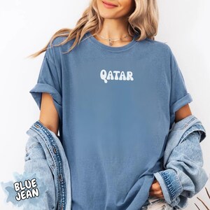 May include: A blue t-shirt with the word "QATAR" in white, stylized text. The shirt has rolled-up sleeves and is paired with a denim jacket. The phrase "BLUE JEAN" is in a blue bubble in the lower left corner.