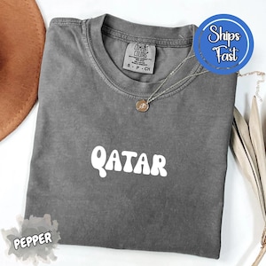 May include: A gray, short-sleeved t-shirt with the word "QATAR" in white, bold letters. The shirt has a crew neck and a tag that reads "COMFORT COLORS". A silver necklace with a pendant is draped on the shirt. The shirt is folded.