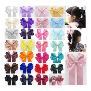 Può includere: Una collezione di fiocchi per capelli in raso colorati, in varie tonalità, tra cui bianco, viola, rosa, giallo e blu. Alcuni fiocchi sono esposti su sfondo bianco, altri sono indossati da una bambina.