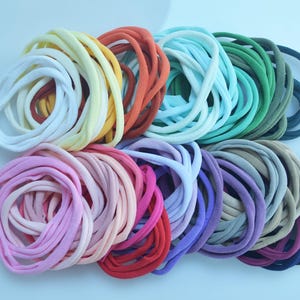 May include: A selection of coiled fabric headbands in a spectrum of colours. The collection includes shades of yellow, orange, blue, green, pink, purple, and black. These headbands are presented in neat bundles, ideal for hair accessories.