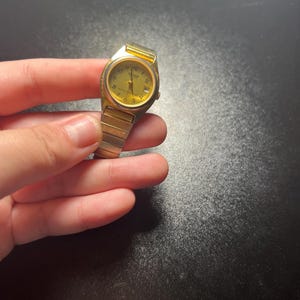 May include: A gold-toned wristwatch with a rectangular face and a matching band. The watch face displays the numbers 1 through 12, with the brand name "SHARP" visible. The watch is held against a dark background.