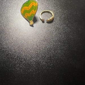 May include: An enamel pin in the shape of a hot air balloon with green and yellow chevron stripes. A gold-colored adjustable ring with a textured snake design is also present.